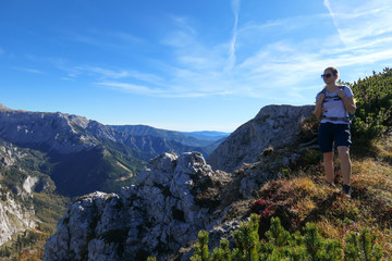 A girl with a hiking backpack standing at the mountain ledge in Hochschwab region in Austria. There are many mountain ranges around her. The slopes overgrown with mountain pine, partially barren rocks