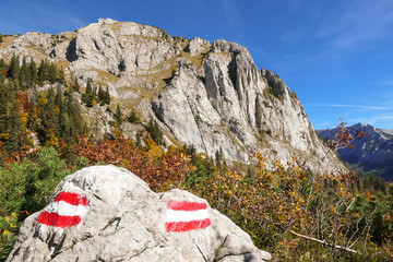A path marks looking like flags of Austria painted on a big stone next to the pathway in Hochschwab region, Austria. There is a massive mountain range in the back. Autumn in the mountains. Gold colors