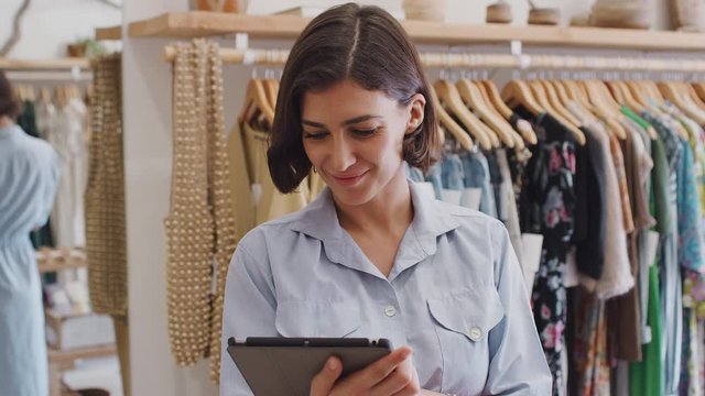 Portrait of female owner of fashion store using digital tablet to check stock on rails in clothing store - shot in slow motion
