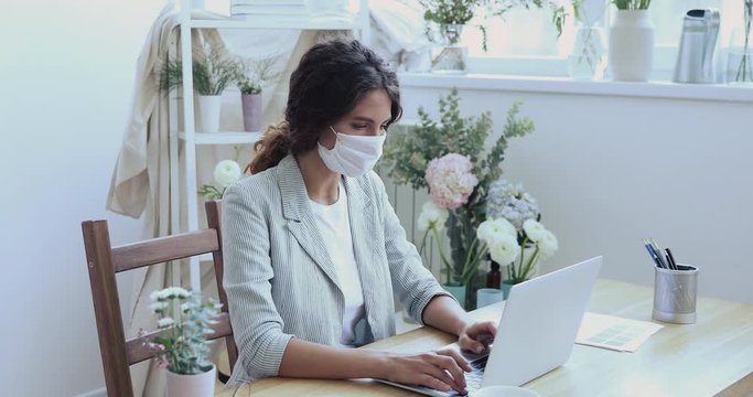 Young employee wears face mask works alone from home office sits at desk using laptop computer. Female designer self-isolating to reduce corona virus spreading. Pandemic quarantine remote job concept.