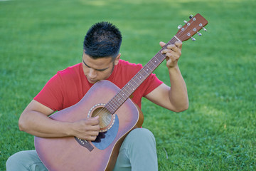 Hispanic male musician playing acoustic guitar standing on green grass and barefoot in a cheerful attitude