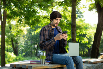 Young freelancer with beard and mustache sitting with a laptop in a park and holding smartphone
