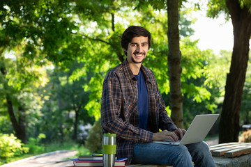 Fototapeta premium Smiling brunet freelancer with beard and mustache sitting with a laptop and coffee in a park looking at the camera