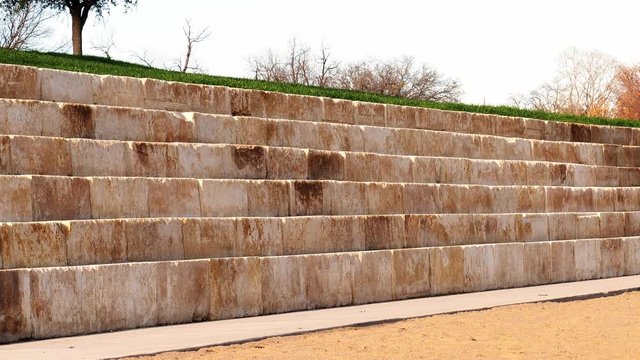 Outdoor amphitheater with large limestone blocks arranged in long rows, designed for seating in a public space, on a sunny afternoon with blue sky.