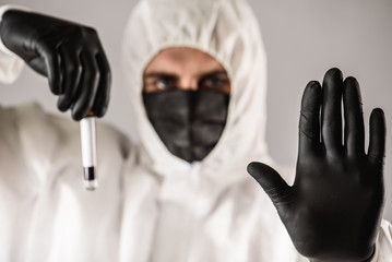a virologist in a black mask and medical gloves wearing a white protective suit, conducts blood...