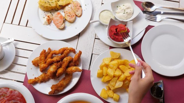 Woman pick up one fried potato cube and dip it into red sauce, eat then. Top view of served table at Spanish cafe outdoor terrace. Tourist try popular local meals and tapas for dinner