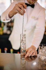 Bartender in a white suite and black bow tie cooling a highball glass with ice cubes mixing them...