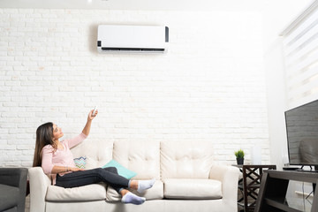 Woman Turning on Air Conditioner At Home - obrazy, fototapety, plakaty