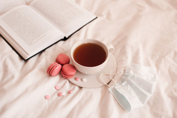 Cup of tea with macaron cookies and meds with medical mask and open book closeup in bed. Good morning. Quarantine at home time. Social isolation.