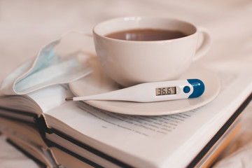 Cup of tea with thermometer with normal temperature and medical mask on stack of book closeup.