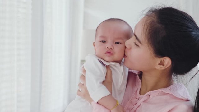 Asian beautiful mother giving warm hugs little baby girl in arms holding softly then kiss young kid in bedroom at home. Soft touch, love care, protection of mom and child in family relationship.