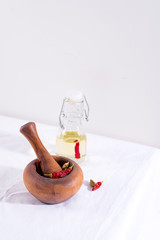 Wooden mortar, pestle and red chili paper in it with bottle of olive oil on the white tablecloth against light grey wall.