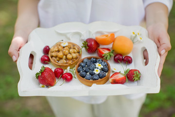 Woman holds white wooden tray with fresh fruits and tarts with berries and nuts