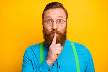 Closeup photo of funny red head guy tricky mood hold finger on lips asking not talk say speak tell wear specs bright blue shirt green suspenders isolated yellow color background