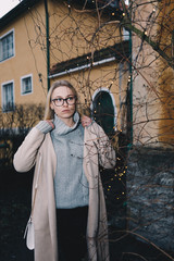 Portrait of long haired blonde woman wearing a sand coat with wool sweater, and black jeans, standing behind old fashioned house. Mystical place with light garland