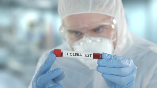 Defocused lab assistant wearing protective suit examines laboratory tube with cholera test