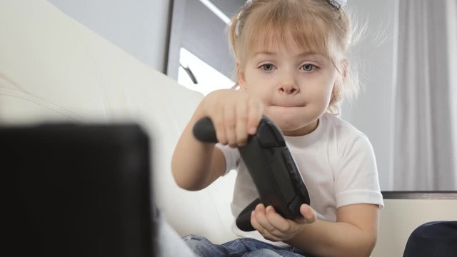 A blonde girl is playing a game console and smiling. A child sitting on a white sofa at home with a joystick in his hands.