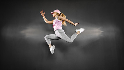 Fototapeta na wymiar Young blonde woman with fit body jumping and running against grey background. Female model in sportswear exercising.