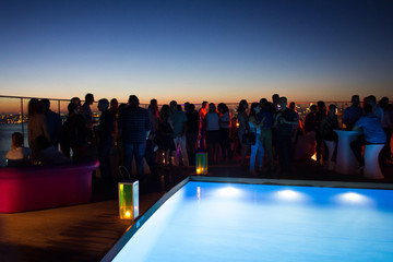 Party crowd of people having fun at roof top club with pool at sunset.