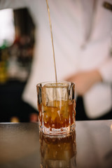 Bartender in a white suite and black bow tie pouring a brown color cocktail from a mixing pitcher into a rocks glass with large ice cube using strainer. Smooth image with shallow depth of field.