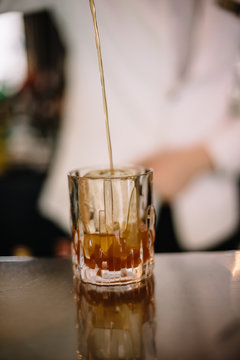 Bartender in a white suite and black bow tie pouring a brown color cocktail from a mixing pitcher into a rocks glass with large ice cube using strainer. Smooth image with shallow depth of field.