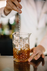 Male bartender in a white suite and black bow tie mixing brown color cocktail in a glass pitcher using long bartenders spoon. Smooth image with shallow depth of field.