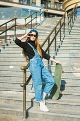 Stylishly dressed woman in blue denim jumpsuit posing with skateboard. Street photo. Portrait of girl holding skateboard. Lifestyle, youth concept. Leisure, hobby and skate in the city