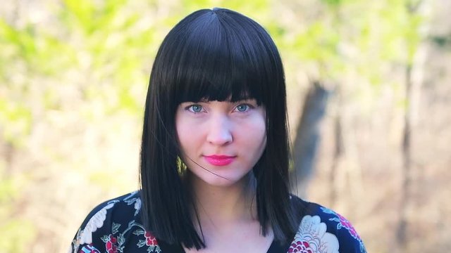 Closeup of young happy Caucasian woman in black hair Asian wig head and shoulders standing outside outdoors in outdoor park garden with colorful trees forest sunny background