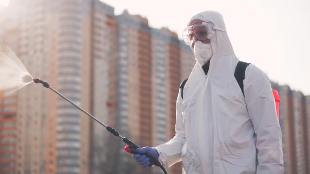 A man in protective equipment disinfects with a sprayer in the city. Surface treatment due to coronavirus covid-19 disease. A man in a white suit disinfects the street with a spray gun. Virus pandemic