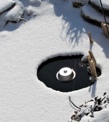 View of a Koi pond covered with snow and an electric pond heater melting the ice 