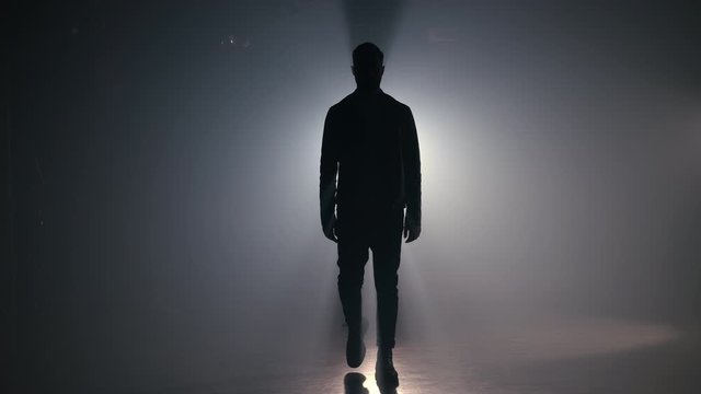 Young man in leather jacket is walking through smoky studio or scene directly to camera. Backlight, bright spotlight on background