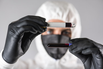A virologist in a medical mask and protective clothing holds a test tube with a blood sample for...
