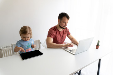 Caucasian man, father of cute baby should work at home becouse of coronavirus and quarantine. He works online in laptop, the child has own laptop.