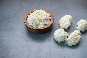 Cauliflower rice on a wooden bowl with some pieces of cauliflower on concrete background.