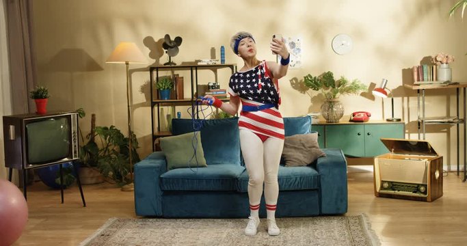 Funny American grey-haired pretty woman standing in room with jump rope while making video on smartphone and talking. Old beautiful retro style female model posing to phone camera at blue sofa indoors
