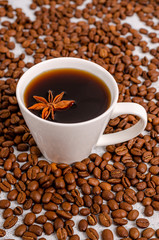 A cup of black aromatic coffee in a white cup with an anise star on a background of roasted arabica coffee beans on a light table.
