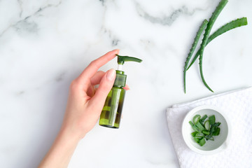 Aloe vera gel green plastic bottle in female hand on marble background with aloe plant stems....