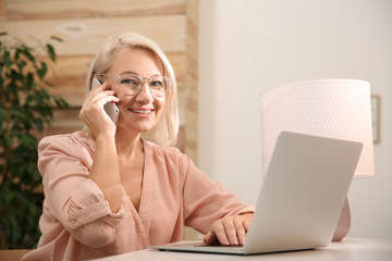 Beautiful mature woman with laptop talking on smartphone at home