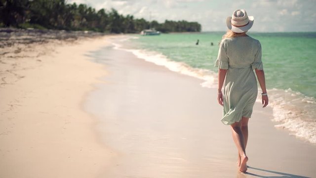 Beautiful Woman Relaxing On Seychelles Flowing Dress Blowing In Wind.Girl Walking Tropical Hawaii Beach.Woman In Hat Walks Along Beach On Caribbean Coast.Travel Mood Wind Blows Dress On Ocean Resort.