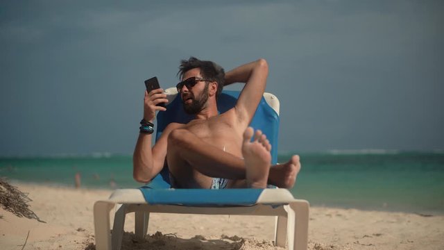 Handsome Guy Scrolling On Smartphone Texting Messages On Social Networks And Lying On  Sunbed.Man Using Mobile Phone On Vacation.Traveller Looking At Cell Phone Using App And Browsing In Internet.