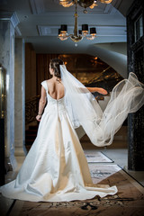 A young bride in a white luxury dress. Woman stands backs to the camera. Her veil flies apart. Magnificent elegant bride on the hotel hall. Black grand piano