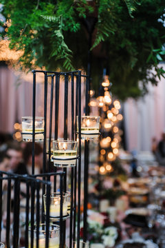 Elegant wedding table centerpiece featuring a tall black metal geometric stand with lit glass votive candles. Lush green ferns hang above. Blurred background shows a festive reception with bokeh light