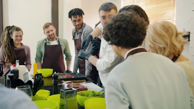 Professional chef is cooking meal teaching students talking enjoying work in kitchen while people in aprons are watching and learning. Youth and food concept.