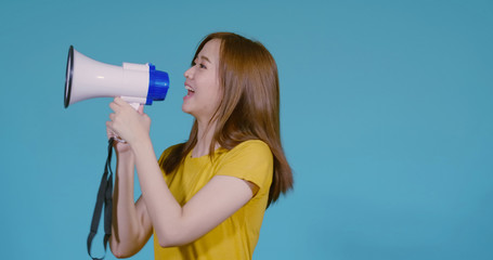Speaking Loud noise announce of young asian woman with megaphone in yellow T-shirt smiling emotion on blue background isolated studio shot