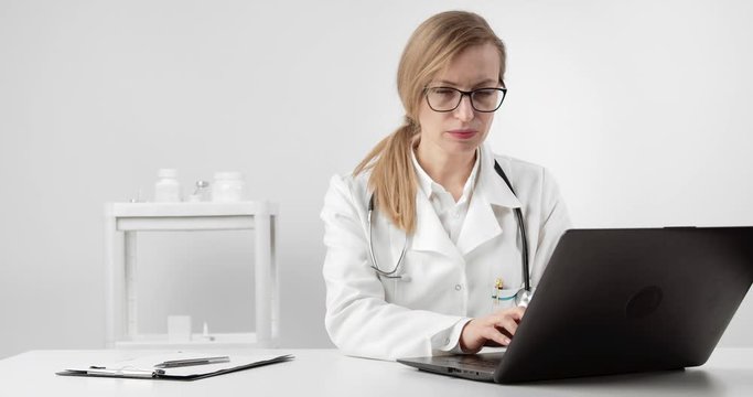 Concentrated mature lady in medical clothing and eyewear sitting at table and working on laptop. Doctor with blond hair filling database on computer.