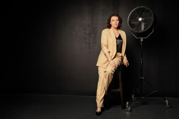 Woman in a yellow suit sits on a chair and looks at the camera on a dark, black background. Free space