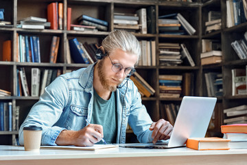 Focused young man student wear headset make notes e study online in library listen lecture internet...