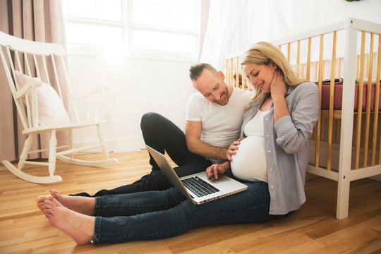 Happy Man And His Pregnant Wife With Computer On The Baby Room At Home