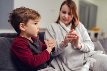 Portrait of small caucasian boy male child kid sitting on the sofa bed at home by mother young woman rubbing hands with alcohol disinfectant antibacterial prevention hygiene product prevent disease