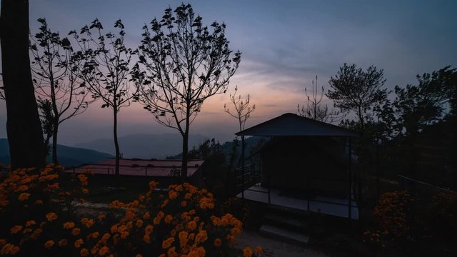 Evening timelapse of sun setting down followed by lights glowing up in a valley with a foreground of Flowers, trees and a tent-house. Wide Shot.